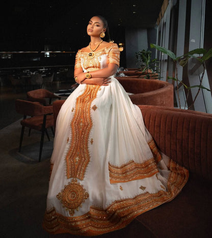 A woman wearing a traditional South Asian ball Ethiopian Store  with white fabric and intricate gold embroidery, posing in a dimly lit indoor setting with velvet seating.