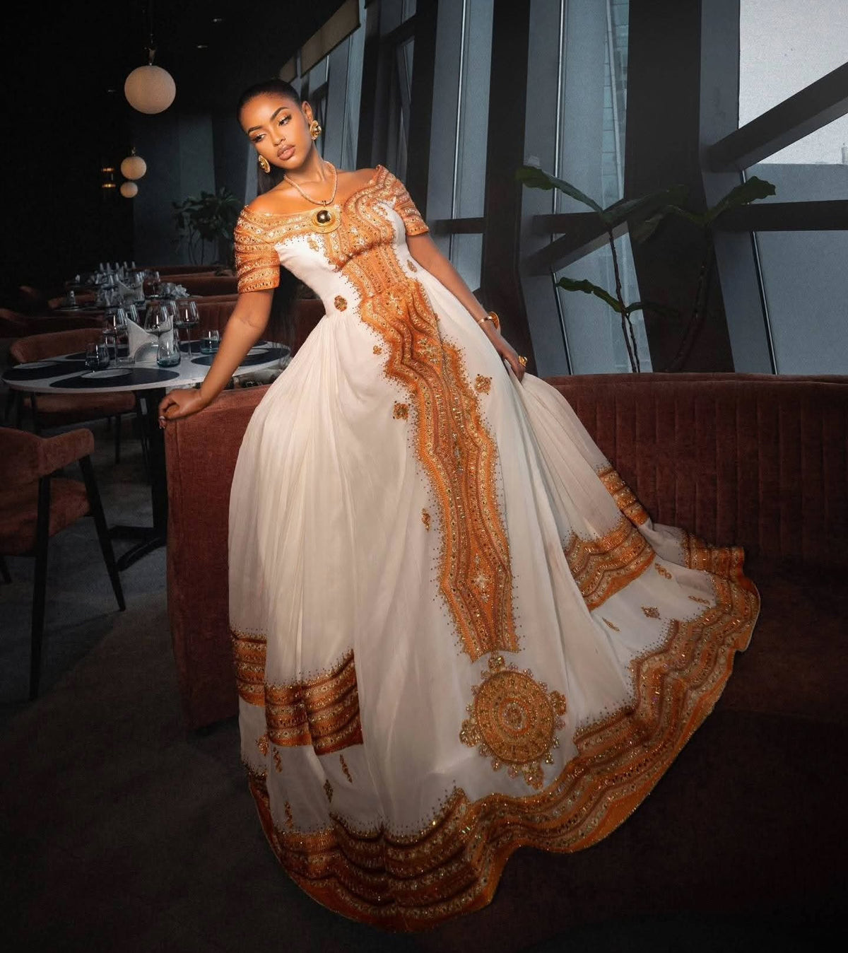 A woman wearing a voluminous off-the-shoulder white Ethiopian Store  with ornate gold embroidery and a wide decorative hem, posed indoors in a dimly lit modern lounge or restaurant setting.