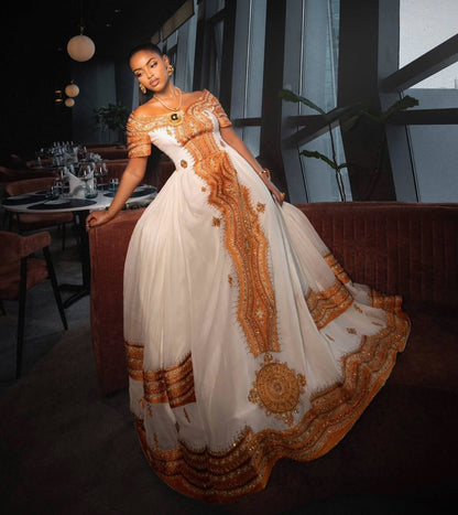 A woman wearing a voluminous off-the-shoulder white Ethiopian Store  with ornate gold embroidery and a wide decorative hem, posed indoors in a dimly lit modern lounge or restaurant setting.