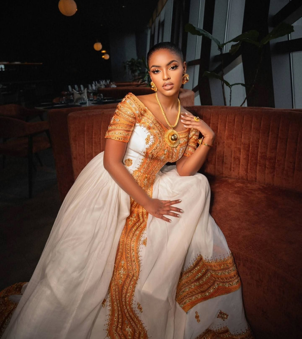 A woman wearing a traditional white Ethiopian Store  with ornate gold embroidery sits on a brown velvet sofa in a dimly lit indoor setting.
