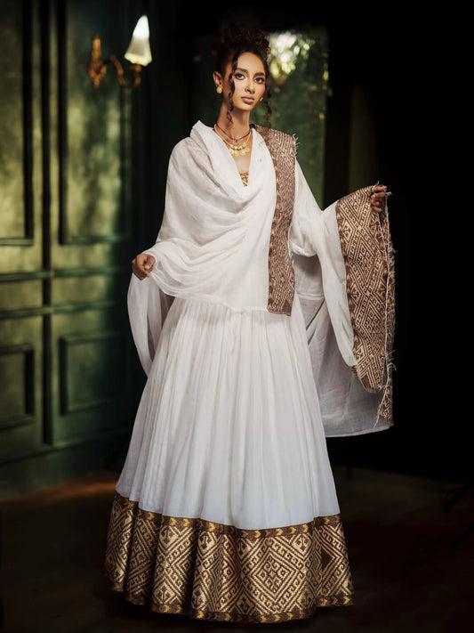 A woman models a traditional white Ethiopian Traditional Clothing with gold patterned borders and a matching draped shawl in a dimly lit interior setting.