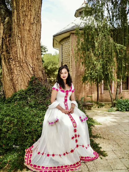 A woman wearing a white traditional Eritrean Zuria with bright pink floral embroidery sits outdoors in a garden courtyard next to a large tree trunk and a brick building.