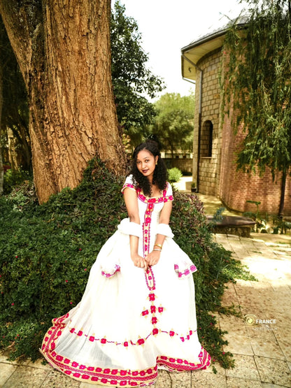 A woman wearing a white traditional Eritrean Zuria with red floral embroidery and a red floral border, posing outdoors next to a large tree and a stone building.