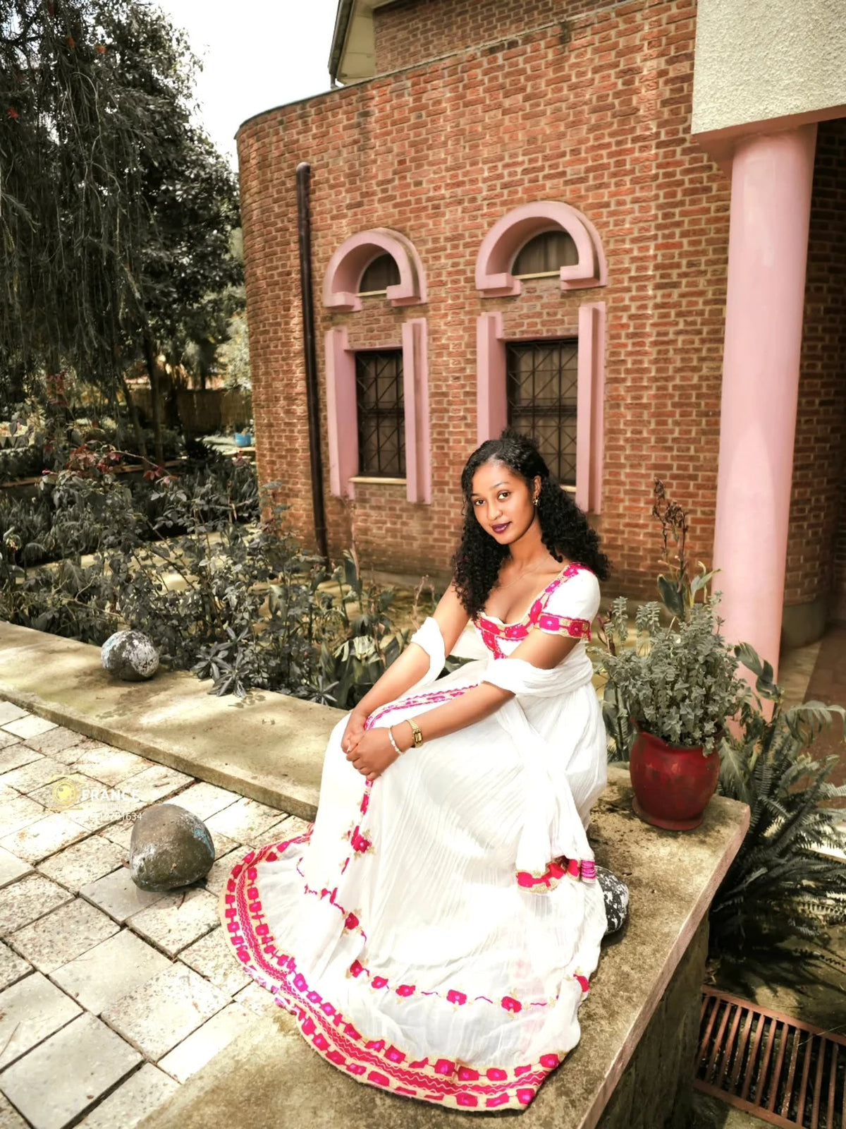 A woman wearing a white traditional Eritrean Zuria with pink floral embroidery sits on a stone ledge in front of a brick building with pink trim.