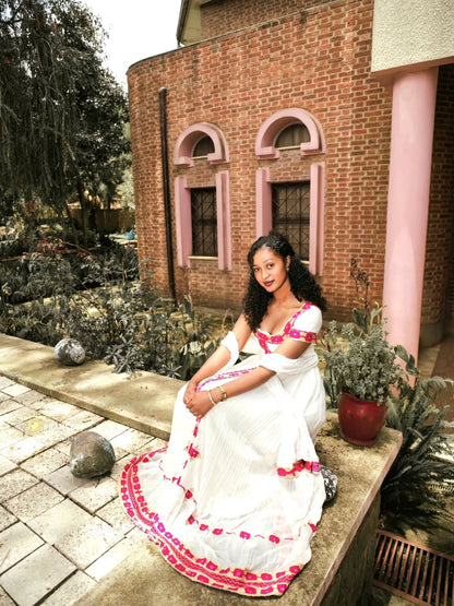 A woman wearing a white traditional Eritrean Zuria with pink floral embroidery sits on a stone ledge in front of a brick building with pink trim.