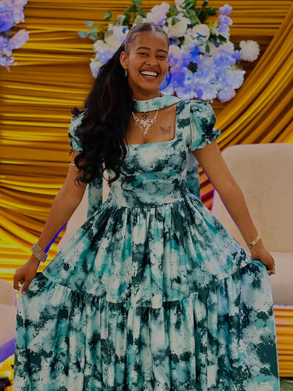 Woman in a green floral Green Habesha Chiffon smiling in front of a decorated background with flowers and yellow drapes.