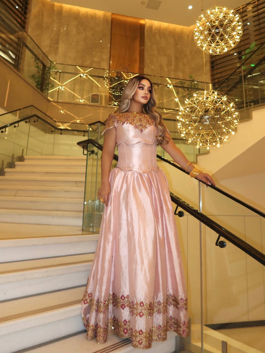 Woman in a pink evening Habesha Kemis  with gold details standing in a luxurious interior setting.