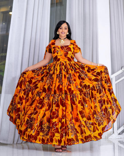 A woman modeling a vibrant, floor-length habesha chiffon with a bold orange, yellow, and brown floral pattern. The dress features cold-shoulder sleeves with tie details, a structured square neckline, and a voluminous, tiered maxi skirt.