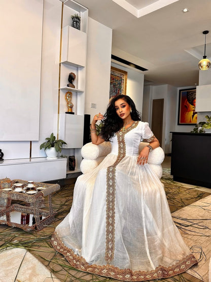 Woman in a white habesha dress with gold details sitting in a modern living room.