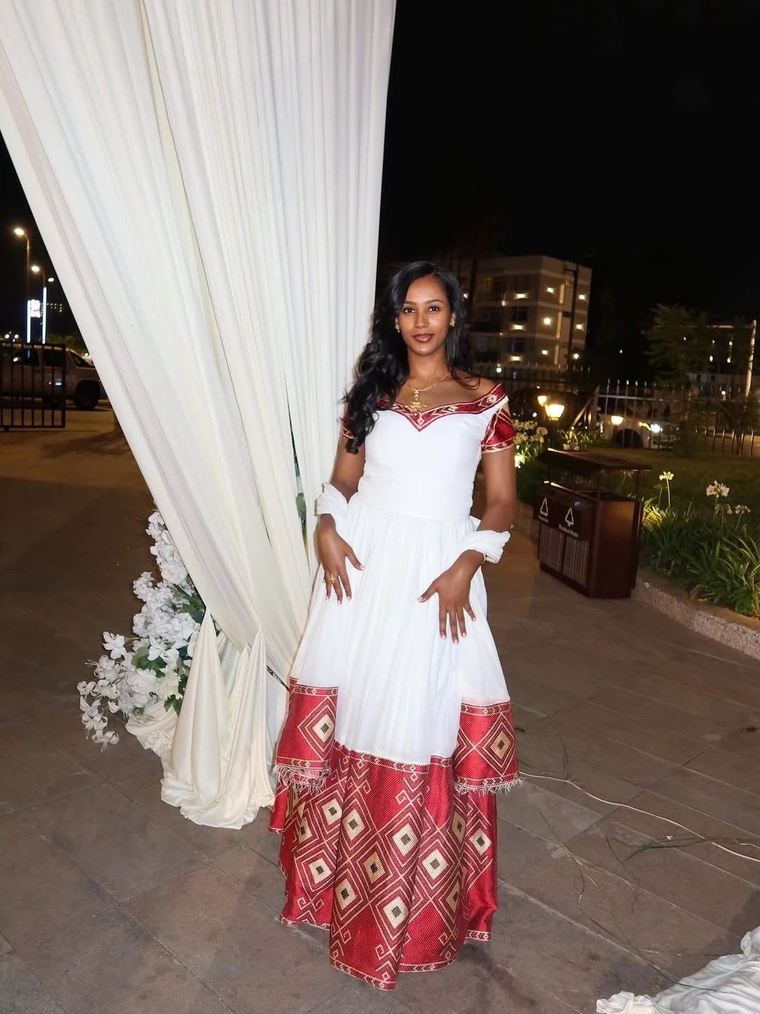 Woman in a white and red Habesha Kemis  standing in front of a white curtain with flowers at night.