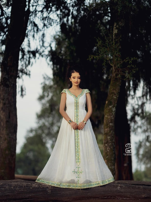 Woman in a white Ethiopian Kemis with green embroidery standing outdoors among trees.