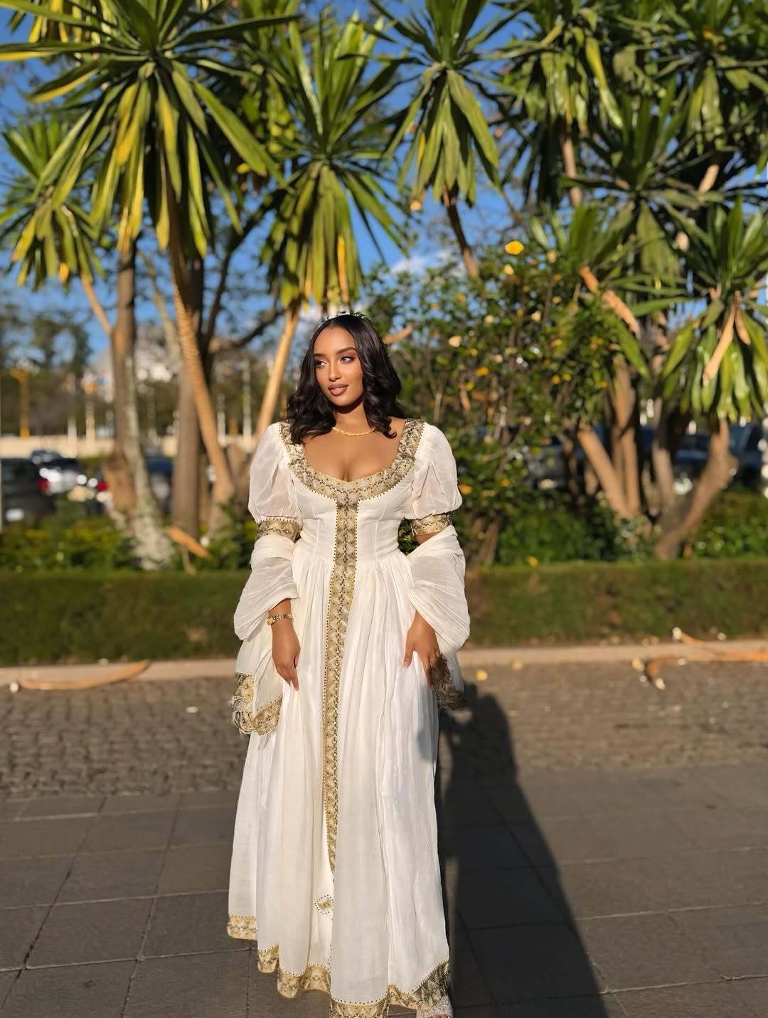 Woman in a white Habesha Zuria with gold details standing outdoors with palm trees in the background