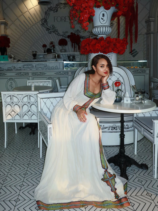 A woman wearing a long white traditional Ethiopian Clothing  with colorful embroidered borders sits at a white table in a decorated indoor setting with patterned tile flooring and white upholstered seating.