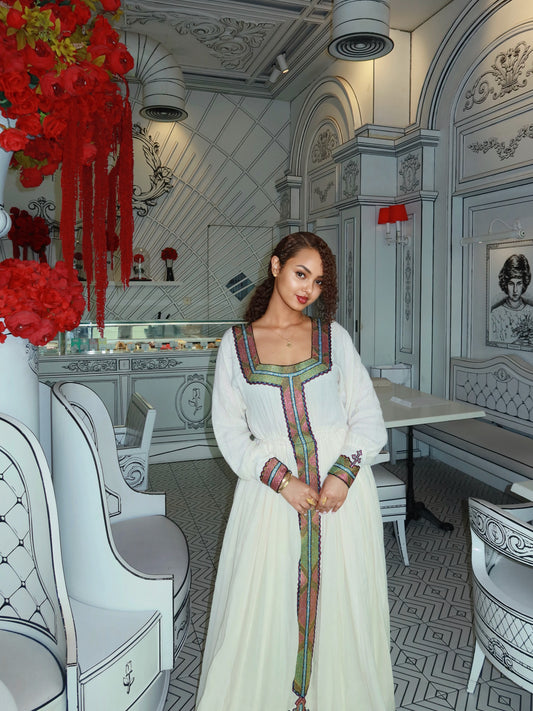 A woman wearing a traditional white long Ethiopian Clothing  with colorful embroidered trim stands in an ornate interior with white chairs, gray patterned floors, and red floral decorations.