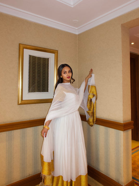 A woman models a traditional white Ethiopian Dresses with a white dupatta and a gold-trimmed skirt, standing in an indoor hallway with beige walls and wood trim.