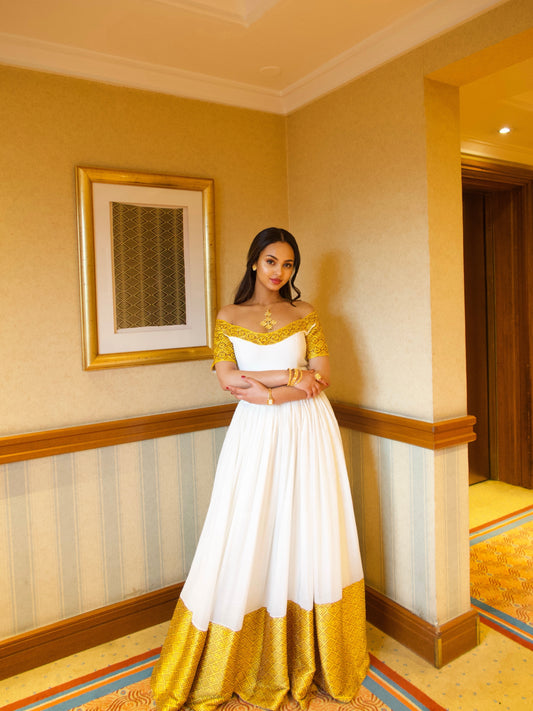 A woman wearing a white and gold off-the-shoulder Ethiopian Dresses stands in a hotel corridor with beige walls and wood trim.