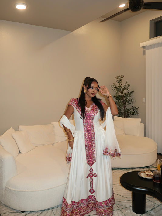 A woman wearing a white traditional Habesha store with pink and red embroidered details stands in a living room in front of a curved white sofa.