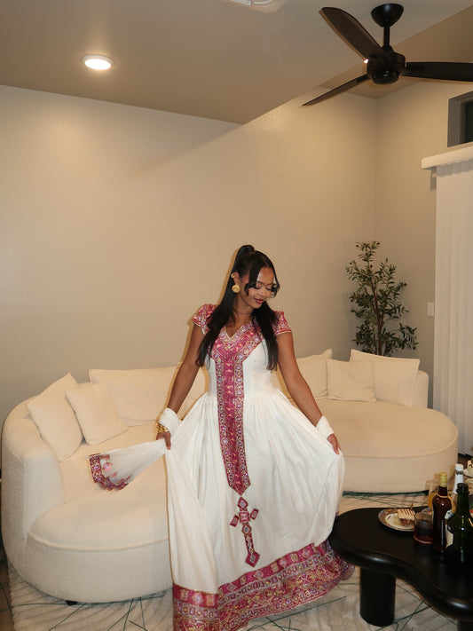 A woman wearing a white traditional Habesha store with pink and red embroidered details stands in a living room in front of a curved white sofa.