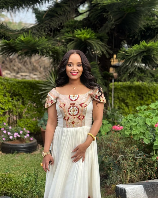 A smiling woman wearing an elegant off-white Habesha Kemis (traditional Ethiopian dress) with intricate red and gold embroidery on the neckline and bodice, standing outdoors in a garden setting.