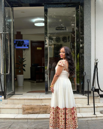 Woman in a white habesha kemis with red and gold patterns standing outside a building entrance.