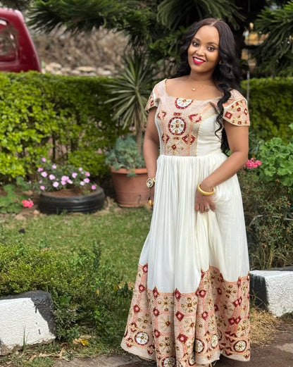 Woman in a white habesha kemis with floral patterns standing outdoors in a garden.