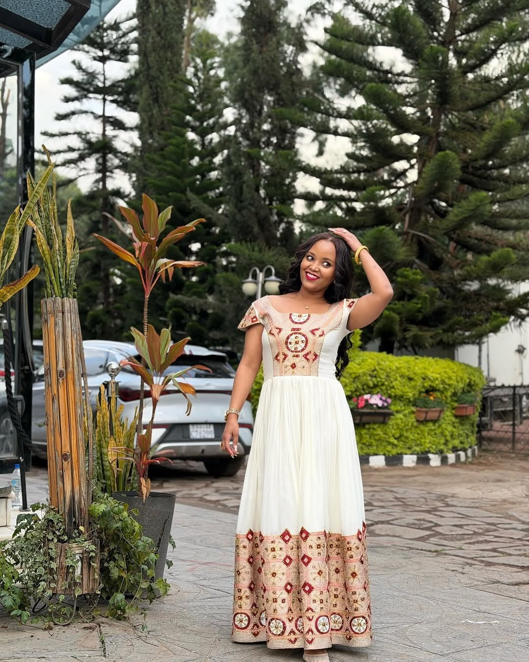 Woman in a white habesha kemis with red patterns standing outdoors near plants and trees.