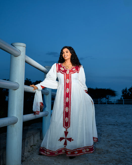 A woman wearing a long white Ethiopian Dress with red and gold embroidery stands outdoors beside a white metal railing at dusk, holding the hem of the dress.