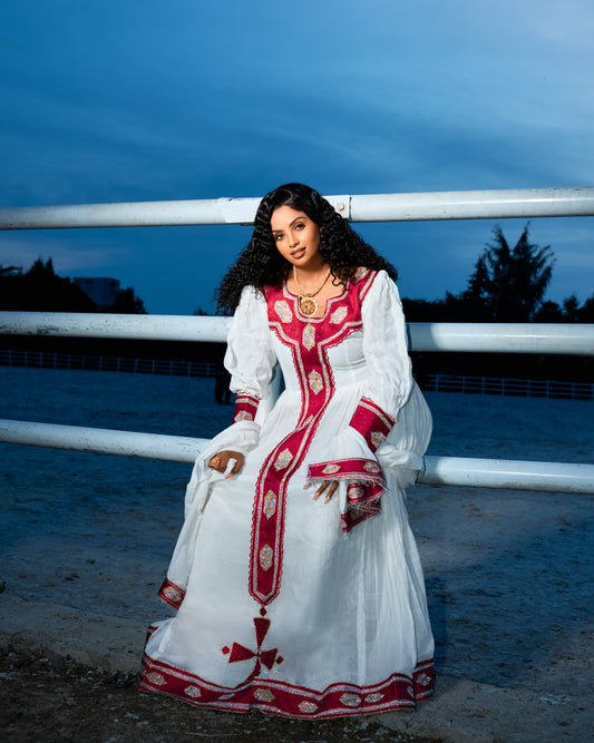 A woman wearing a traditional white Ethiopian Dress with red and gold embroidery, posing outdoors in front of a white railing at dusk.