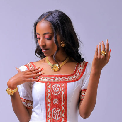 A woman wearing a white Habesha dress  with red and gold embroidered patterns, styled with gold jewelry, including a necklace, earrings, and rings, against a solid light purple background.