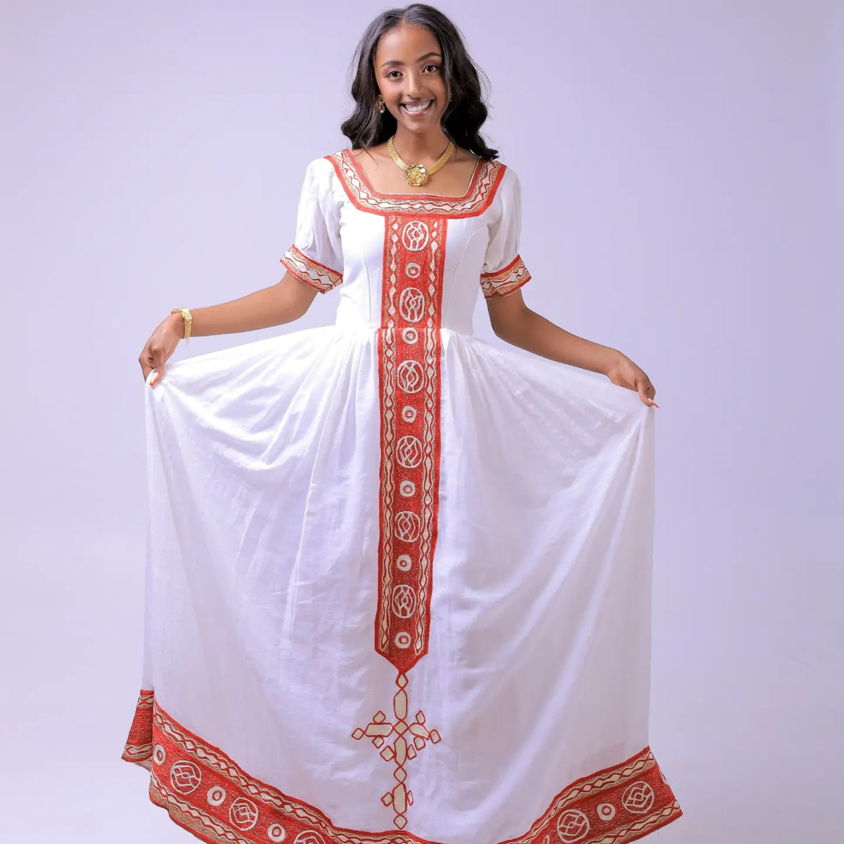 A woman modeling a white traditional Habesha dress  with red and gold embroidered details, holding out the skirt against a light lavender studio background.