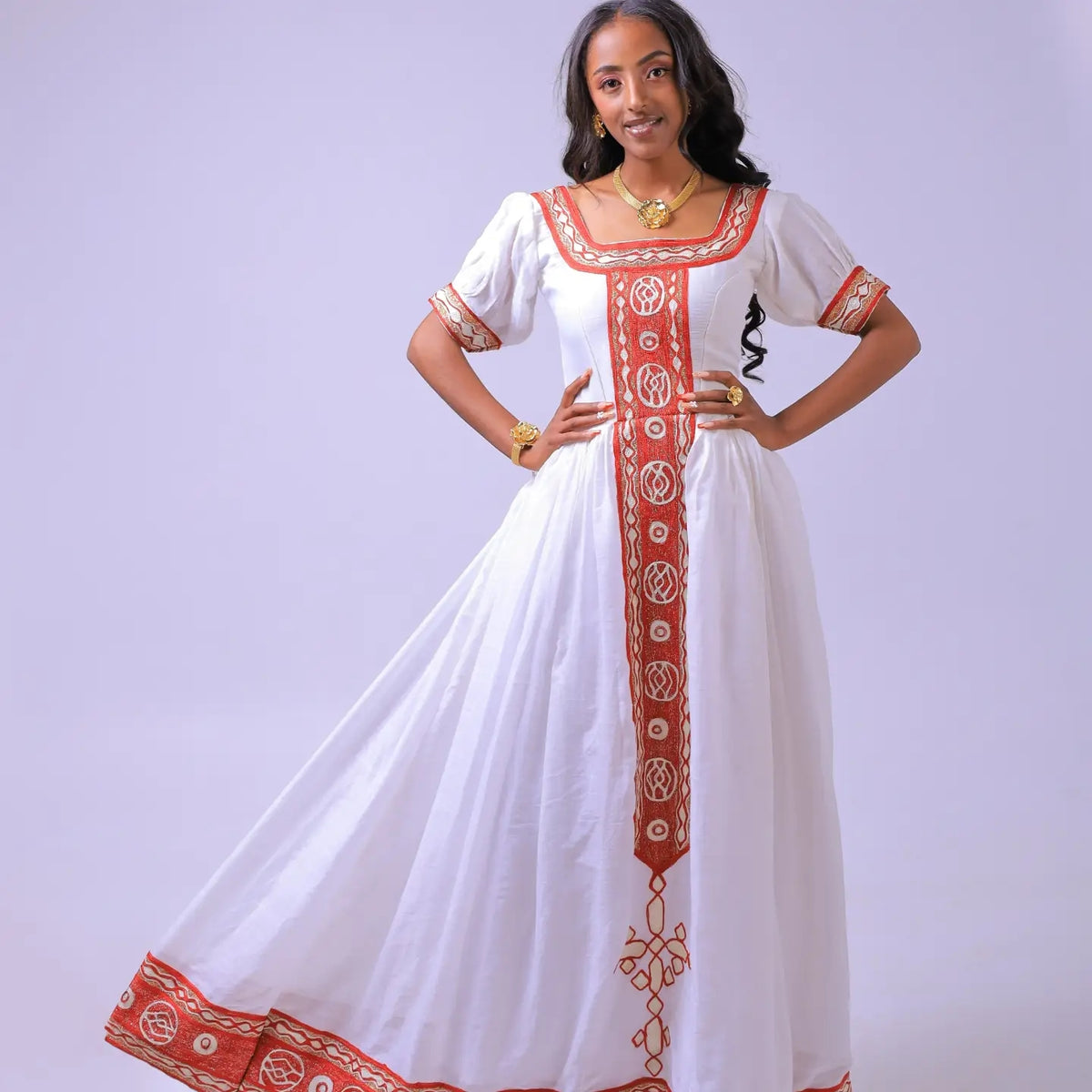 A woman modeling a white maxi Habesha dress  with red and gold embroidered patterns, standing against a light lavender studio background.