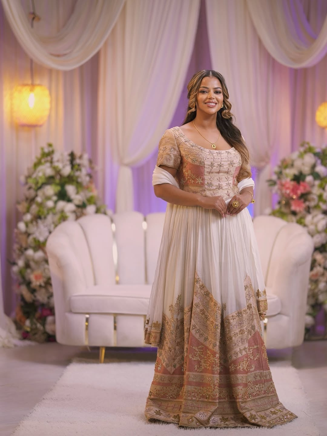 Woman in a traditional outfit standing in a decorated room with floral arrangements and white furniture.