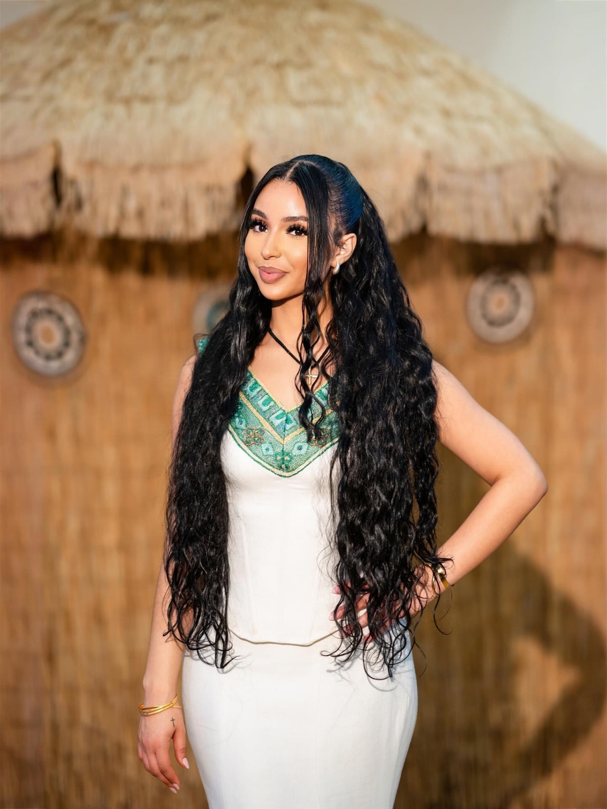 A woman with very long, wavy black hair wearing a white Green Habesha Family matching set with a green patterned neckline, standing in front of a thatched hut background.