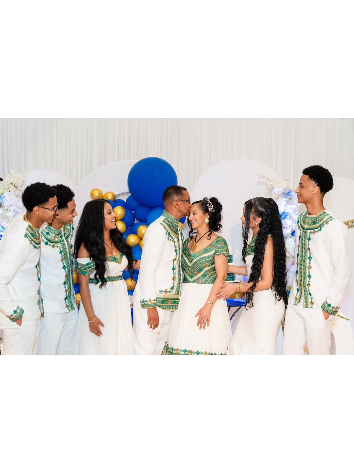 A group of seven people wearing coordinated white traditional-style Green Habesha Family matching set with green embroidered details, standing together in front of a white backdrop with blue and gold balloons.