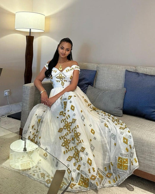 A woman wearing a white off-the-shoulder ball Habesha dress  with gold and gray geometric patterns, sitting on a gray sofa in a living room setting.