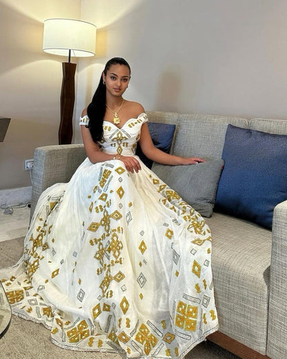 A woman wearing a white off-the-shoulder ball Habesha dress  with gold and gray geometric patterns, sitting on a gray sofa in a living room setting.