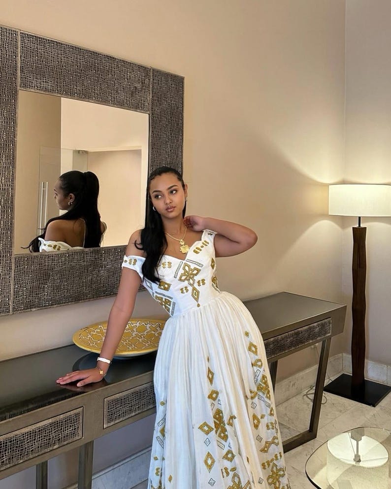 A woman wearing a white off-the-shoulder maxi Habesha dress  with gold geometric patterns, posing in a modern room with a large textured mirror and a floor lamp.
