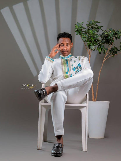Man in a white Habesha Men’s Cultural Shirt  sitting on a chair with a plant in the background