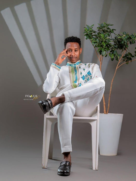 Man in a white Habesha Men’s Cultural Shirt  sitting on a chair with a plant in the background