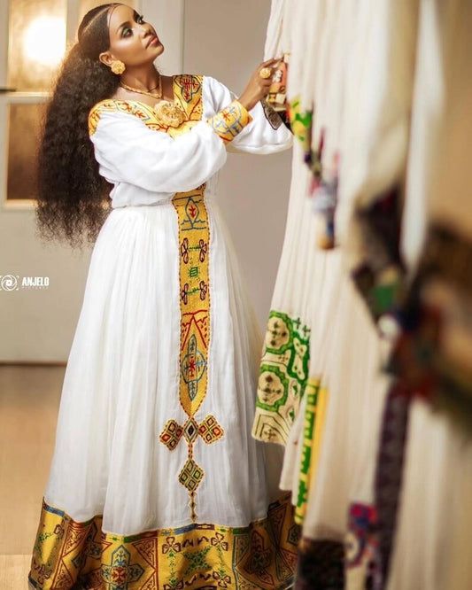 Woman in a Yellow Habesha Dress with colorful patterns, standing in a room.