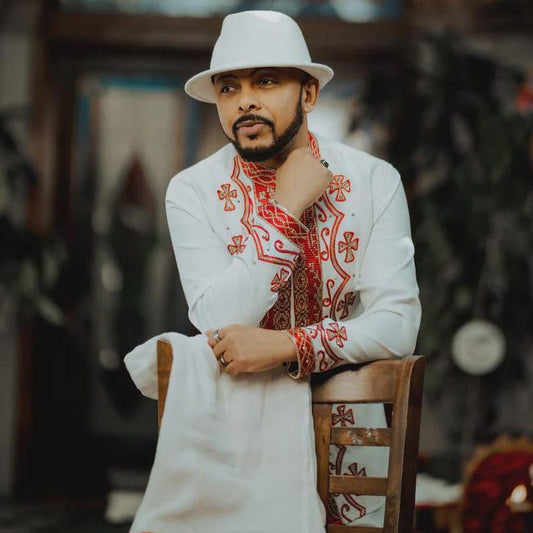 Man wearing a white embroidered Ethiopian Cultural Men’s Shirt and hat, sitting on a wooden chair.