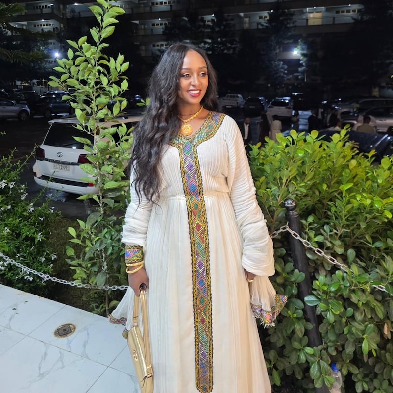 A woman wearing a long cream-colored traditional habesha dress with colorful embroidered details down the front, standing outdoors at night in front of green bushes and parked cars.