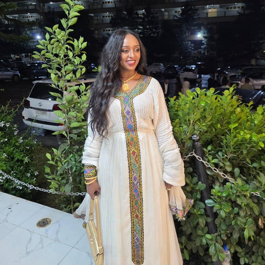 A woman wearing a long cream-colored traditional habesha dress with colorful embroidered details down the front, standing outdoors at night in front of green bushes and parked cars.