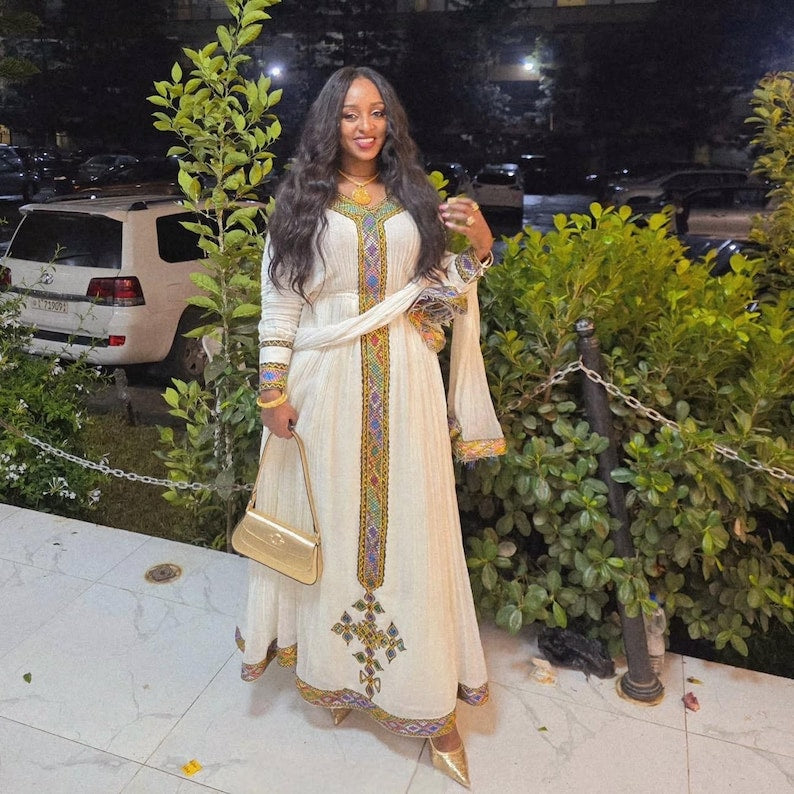 A woman wearing a long cream-colored traditional habesha dress with colorful embroidered details down the front, standing outdoors at night in front of green bushes and parked cars.