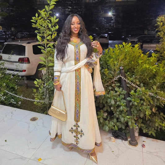 A woman wearing a long cream-colored traditional habesha dress with colorful embroidered details down the front, standing outdoors at night in front of green bushes and parked cars.