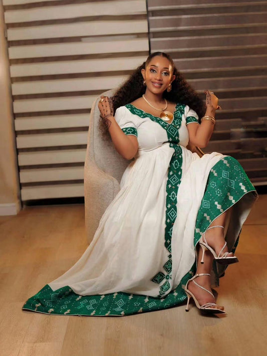A woman wearing a white and green traditional Habesha kemis  with geometric patterns, sitting in a beige armchair in an indoor setting with wooden floors and slatted wall panels.