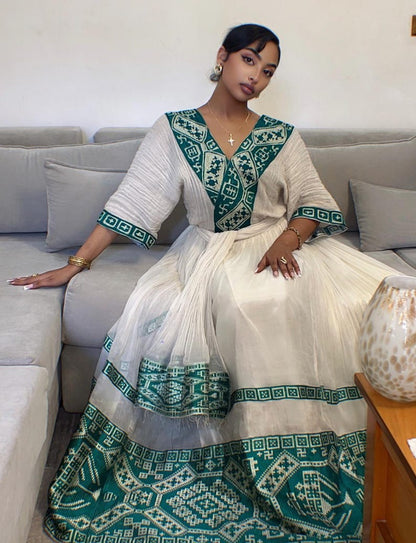 A woman wearing a traditional Indian Habesha dress sits on a gray sofa. The dress is off-white with teal green geometric patterns on the neckline, sleeve cuffs, hem, and lower skirt.