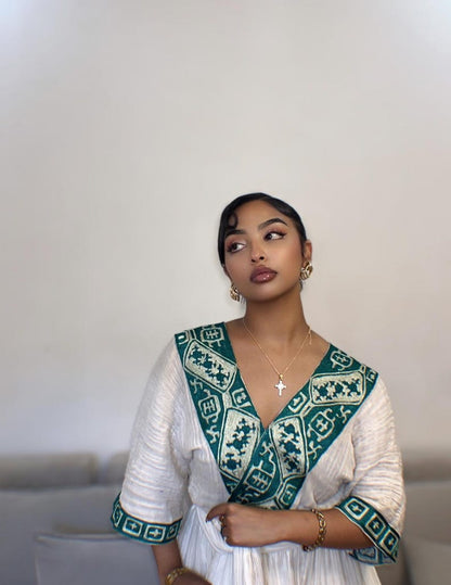 A woman wearing a white textured Habesha dress with a deep V-neckline and wide sleeves, featuring a prominent teal and cream geometric embroidered border along the neckline and cuffs. She is wearing gold jewelry, including a cross necklace and hoop earrings, and is posed against a neutral off-white wall.