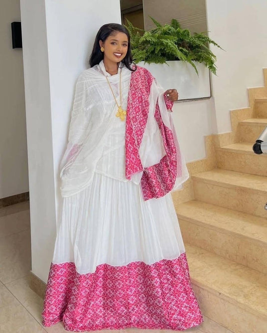 A woman modeling a traditional white maxi Habesha kemis with a vibrant pink and white floral border, paired with a matching sheer white shawl draped over her shoulders, set in an indoor environment with beige stone stairs.
