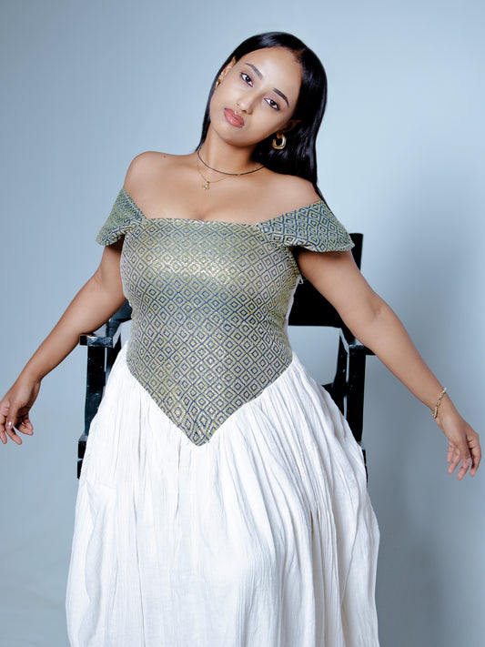 A woman posing in a white maxi Habesha Dress with an off-the-shoulder, geometric-patterned bodice, seated on a black chair against a light gray background.